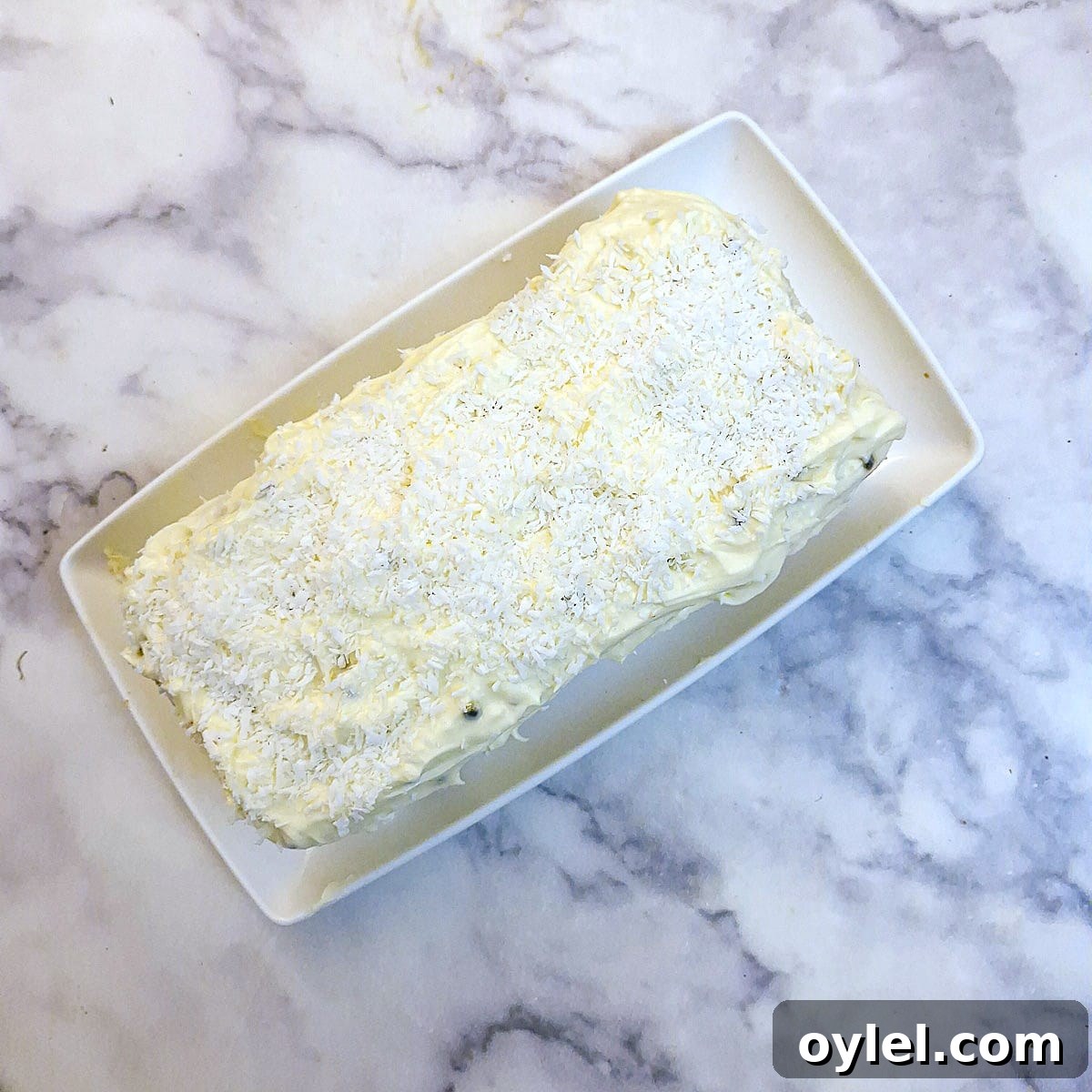 The frosted coconut loaf cake sprinkled with dessicated coconut on a white serving plate.