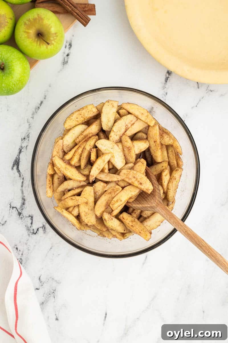 A bowl of seasoned apple slices with a wooden spoon, next to green apples and a pie crust on a marble countertop.
