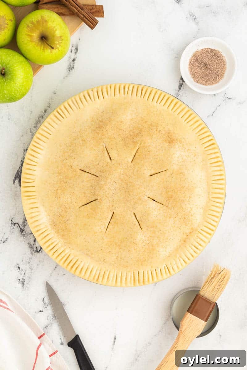 Unbaked apple pie with vent slits on top covered in dough, surrounded by green apples, a bowl of sugar and cinnamon, a knife, a pastry brush, and a cloth on a marble surface.