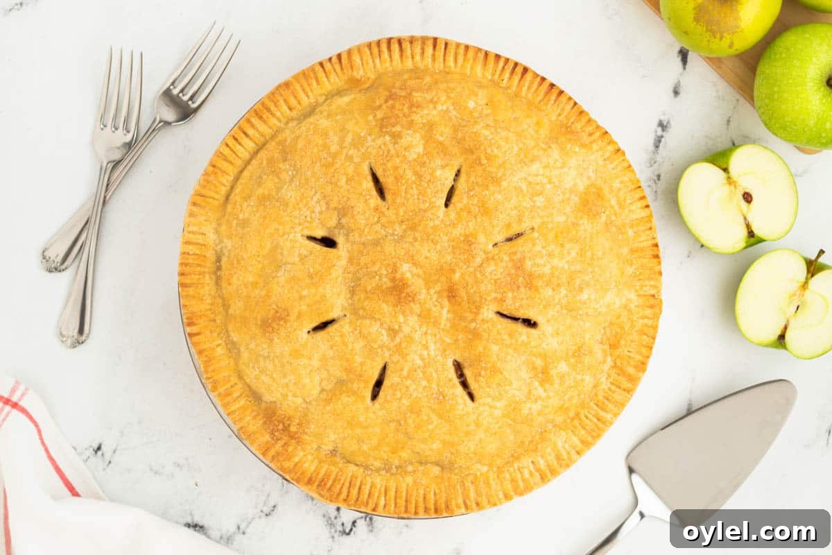 A golden-brown baked pie with a crimped edge sits on a marble surface. Three forks, a pie server, and a napkin are to the left, with sliced green apples on a cutting board to the right.