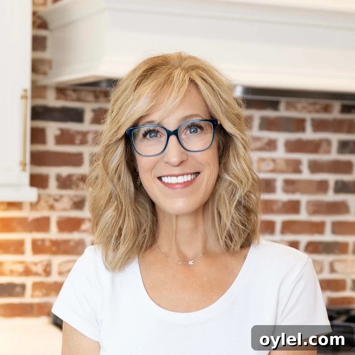 Person with shoulder-length blond hair and blue glasses smiling indoors in front of a red brick wall.