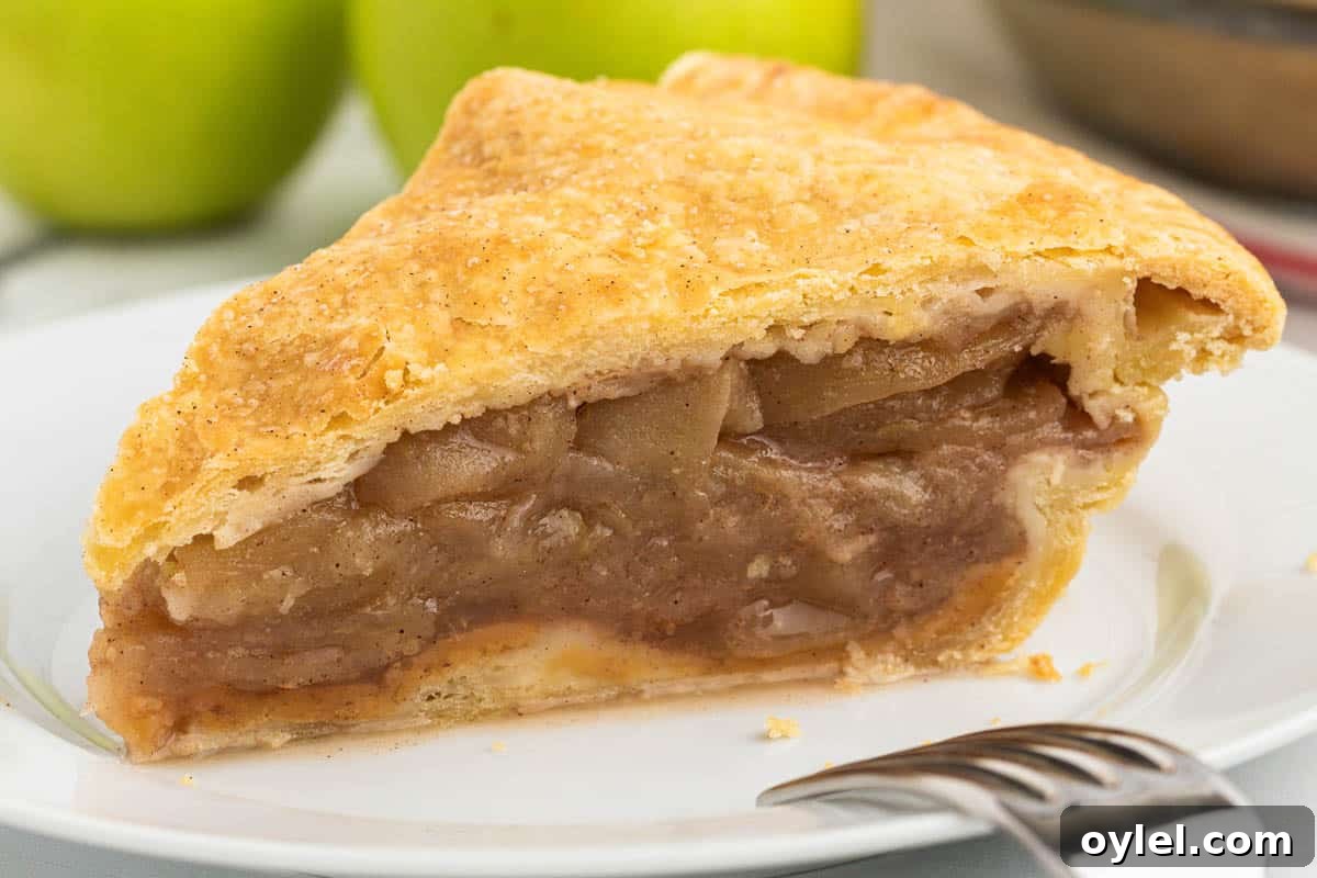 A slice of apple pie with a golden crust, filled with seasoned apple chunks, is served on a white plate next to a fork. In the background, there are green apples.