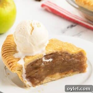 A slice of double crust apple pie with a scoop of vanilla ice cream on top, served on a white plate. An apple and a striped cloth are in the background.