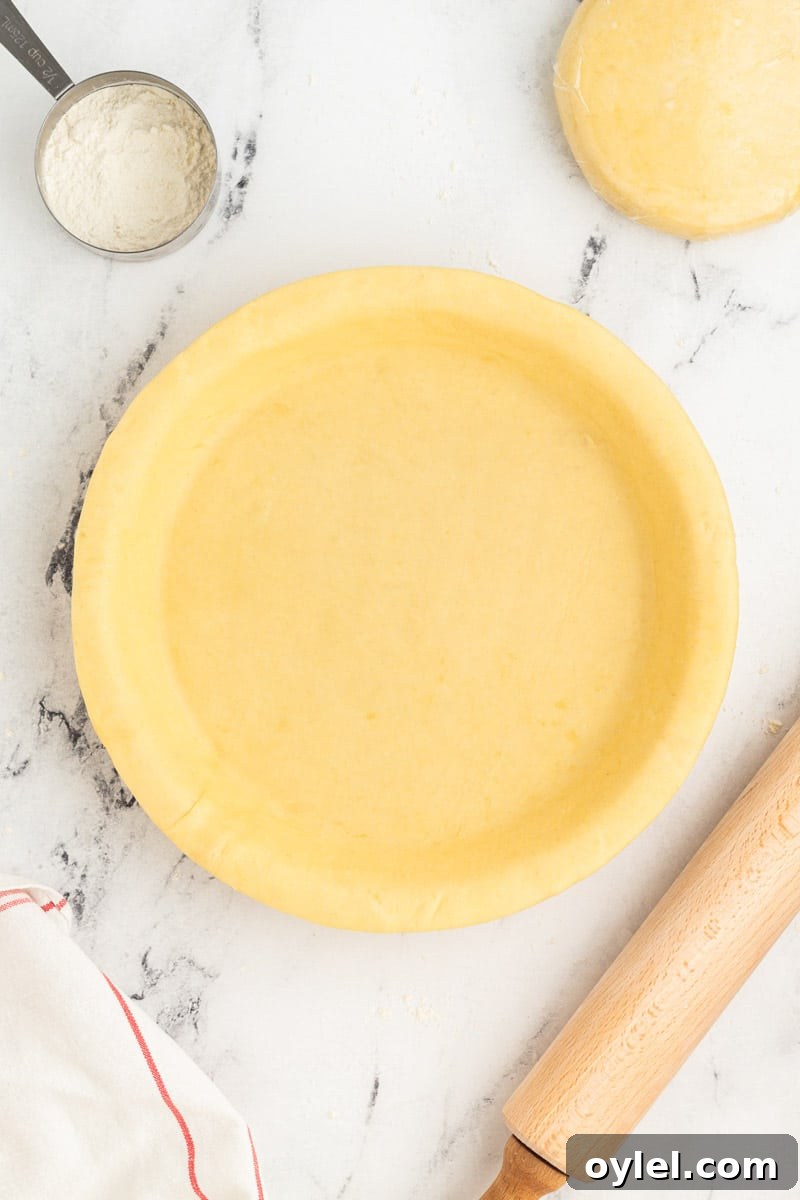A round pie crust is neatly placed in a pie dish on a marble surface. Nearby are a rolling pin, a measuring cup with flour, and a ball of unused dough.