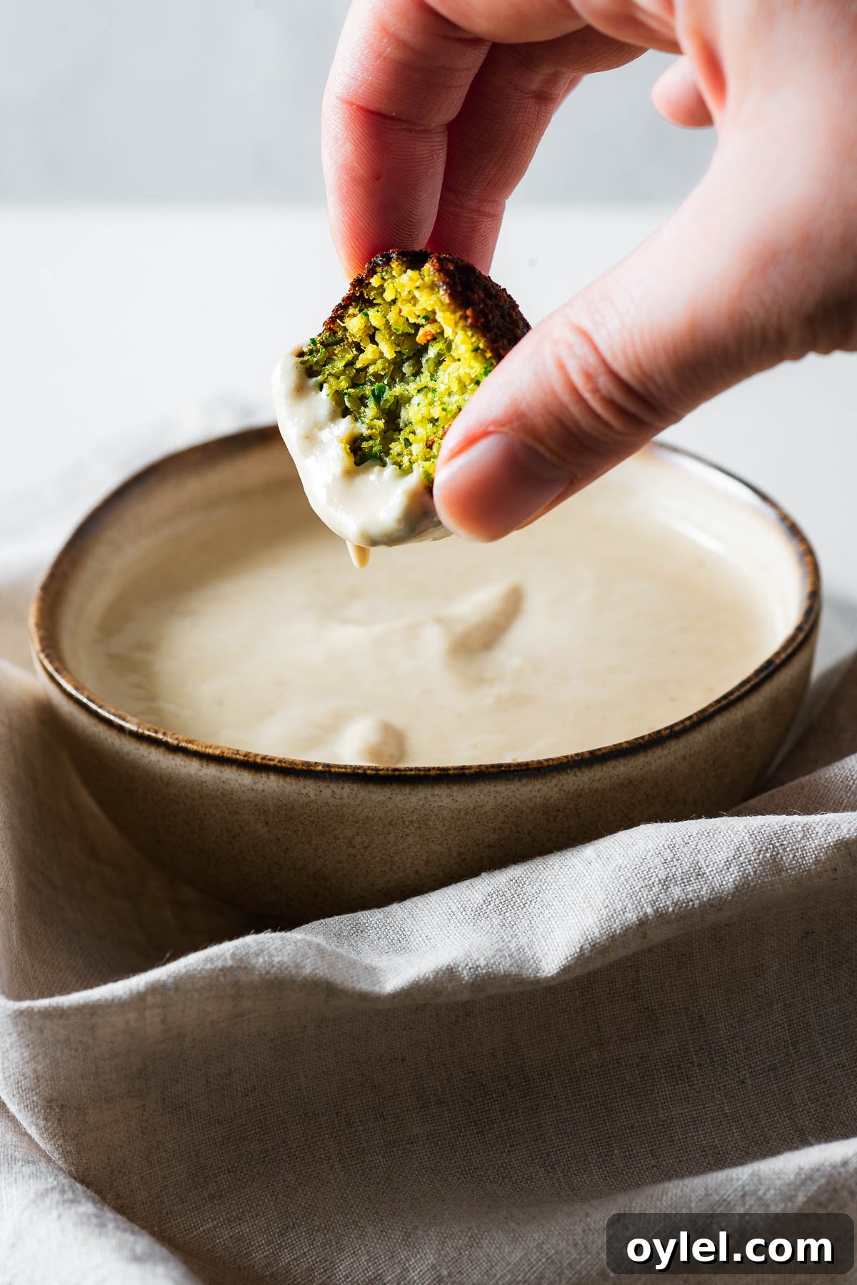 Lemon Tahini Sauce Recipe: 5 Flavor Variations 7 A close-up of a hand dipping a fresh Middle Eastern falafel into a small bowl of tahini sauce.