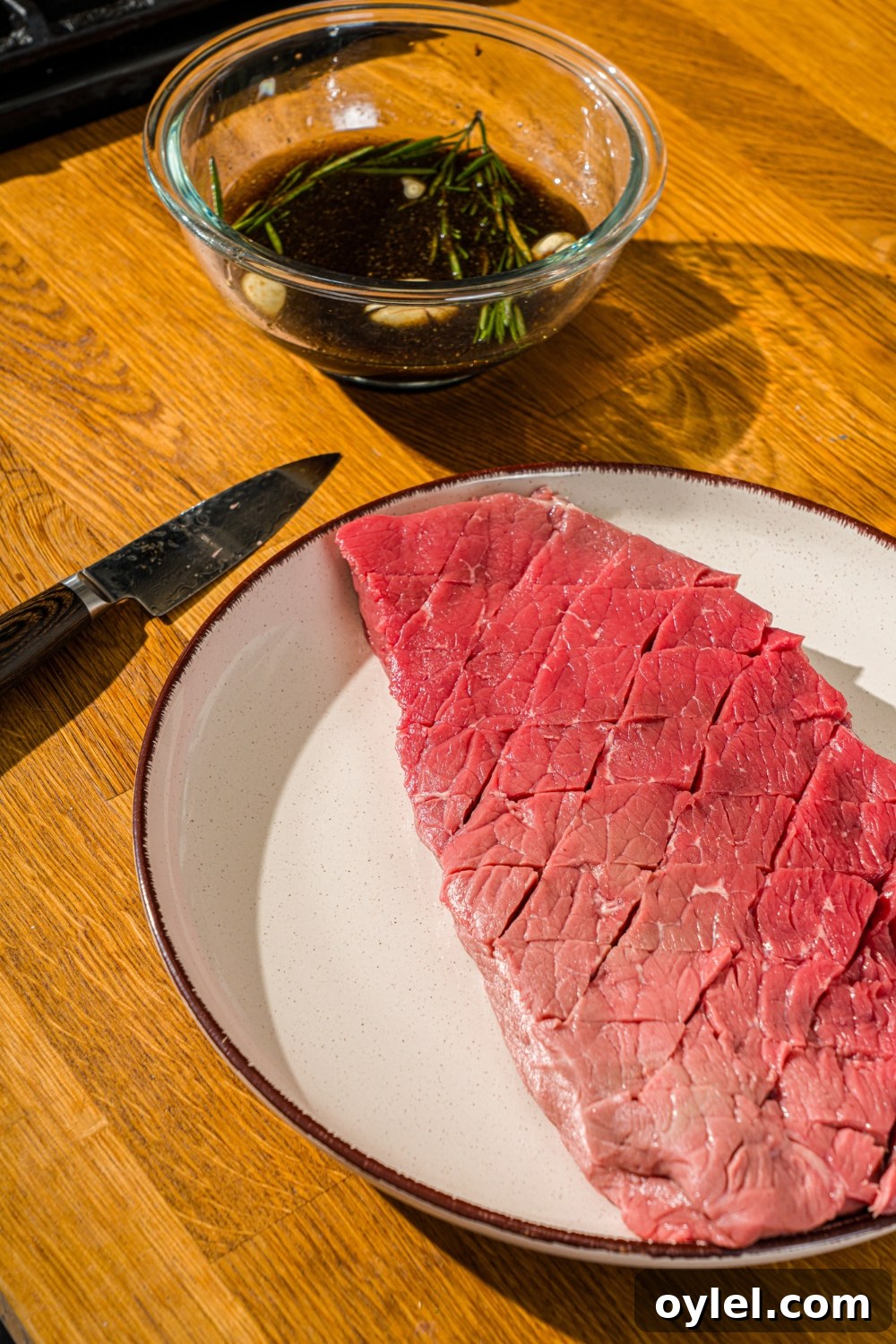 London Broil Marinade Recipe for Tender, Flavorful Steak 6 A white plate with London broil beef scored in a diagonal pattern. The plate is on a wooden counter with a bowl of marinade.
