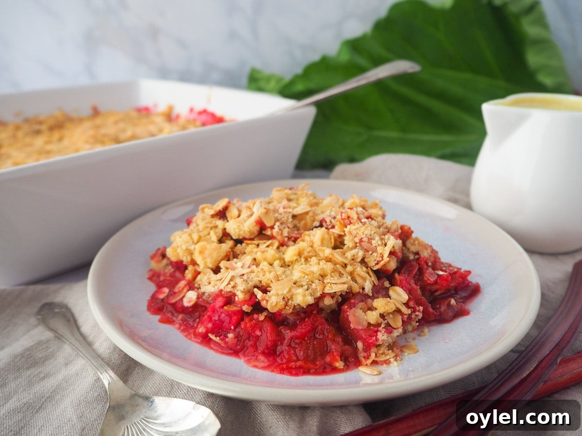 Classic Rhubarb Crumble Recipe 12 serving rhubarb crumble with custard and fresh rhubarb, baking dish of crumble and jug of custard on the side.