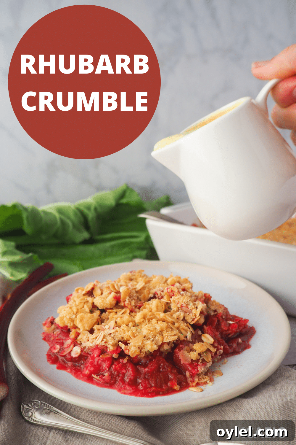 Classic Rhubarb Crumble Recipe 13 Serving of rhubarb crumble with jug of custard, with fresh rhubarb and baking dish in the background, with text overlay, rhubarb crumble and KCAEIC.