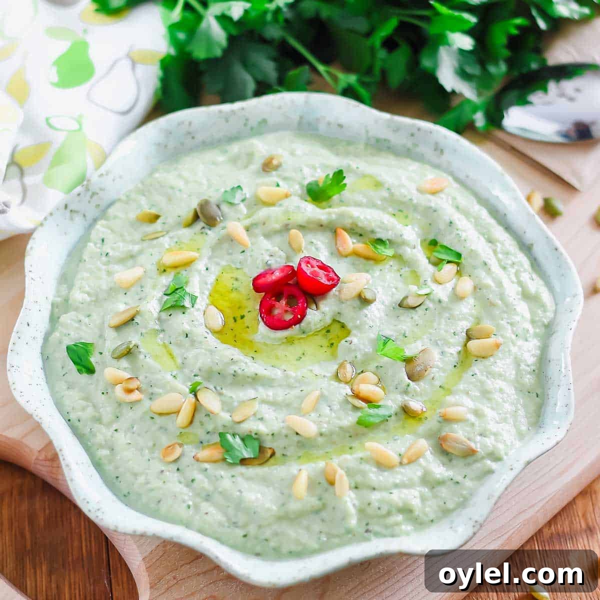 A bowl of roasted eggplant dip garnished with pine nuts, pumpkin seeds, and parsley. Three slices of red chili pepper are at the center. Fresh parsley and a patterned cloth are in the background.