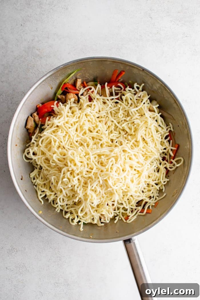Yaki soba noodles added to stir frying chicken, shredded carrots, bell pepper, onions, mushrooms, cabbage, and green onions in a large wok.