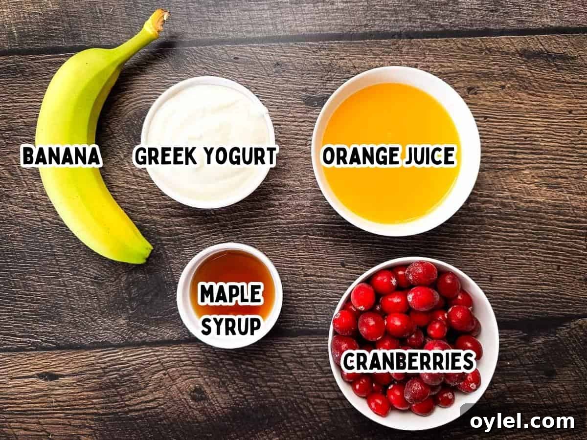 Cranberry Smoothie Recipe with Banana and Yogurt 2 Five simple ingredients laid out on a wooden surface: a banana, a bowl of Greek yogurt, a bowl of orange juice, a small dish of maple syrup, and a bowl of fresh cranberries.