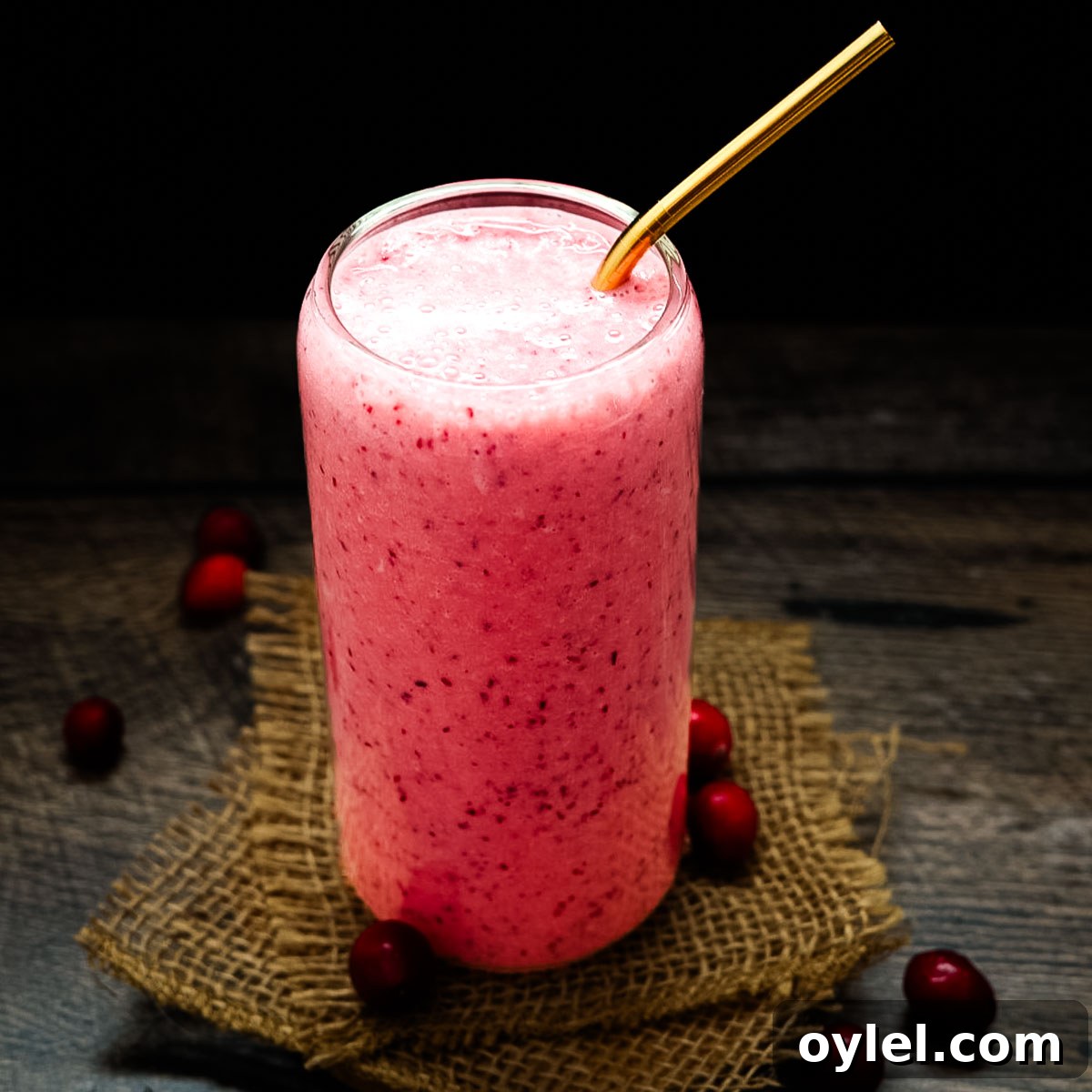 Cranberry Smoothie Recipe with Banana and Yogurt 7 Cold and creamy cranberry smoothie in a clear glass with a gold straw, photographed on a dark wood table with cranberries around the base.