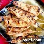 Overhead image of skewers of grilled chicken piled on a square black and gold serving dish on a red cloth.