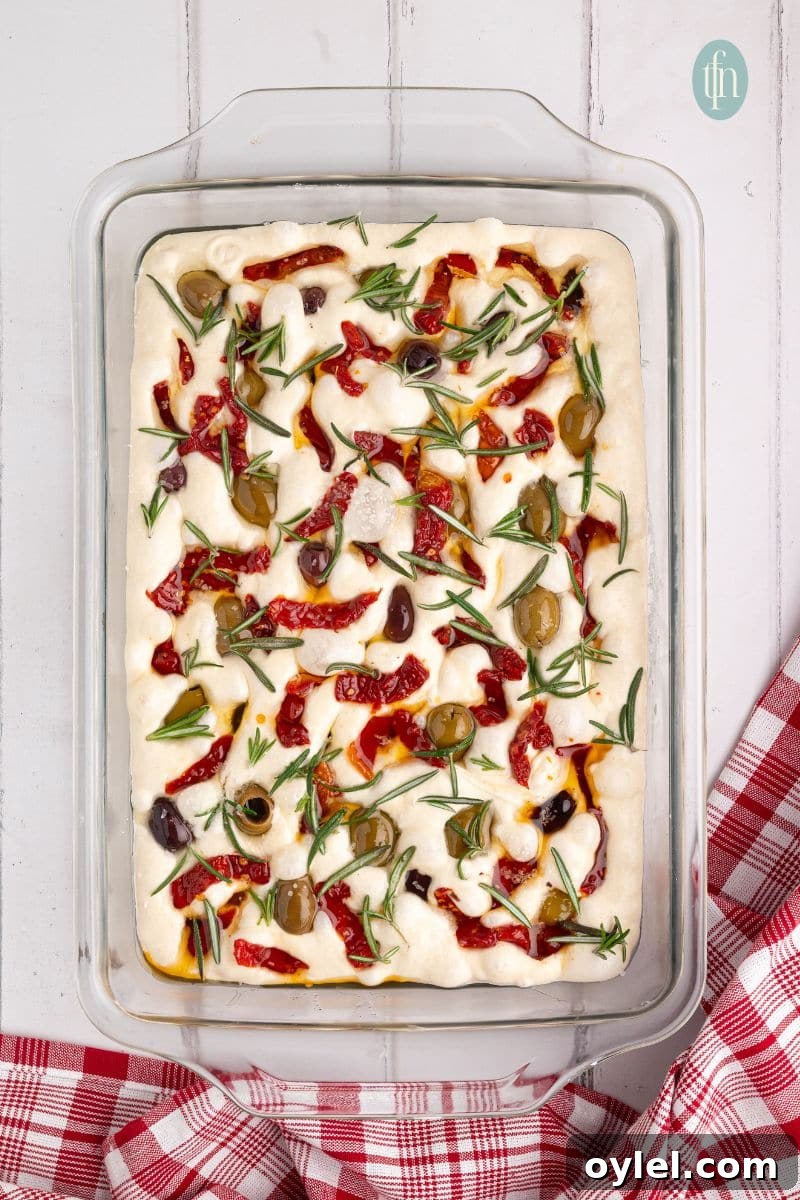Sourdough Focaccia Recipe: Crispy Crust, Airy Interior 8 A dough mixture topped with raw ingredients in a glass pan.