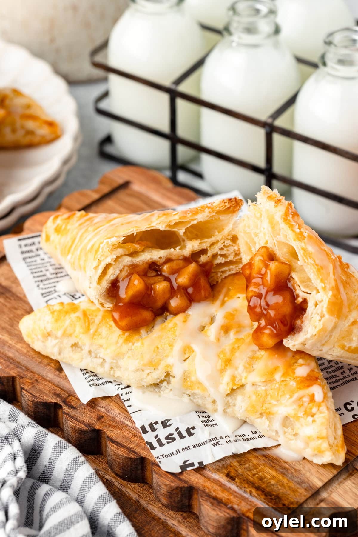 Flaky Puff Pastry Apple Turnovers Recipe 7 Two iced apple turnovers on parchment atop a wooden board, with milk bottles in the background.
