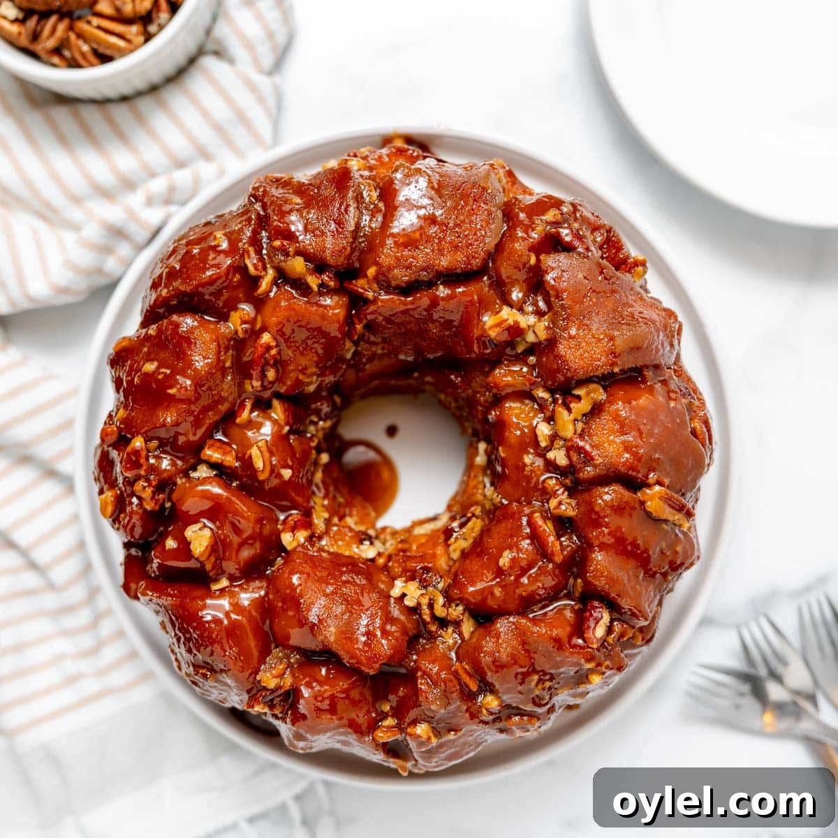 Cinnamon Pull Apart Monkey Bread with Biscuits 2 A round monkey bread loaf topped with caramel glaze and chopped pecans sits on a white plate, with forks and a bowl of pecans nearby.