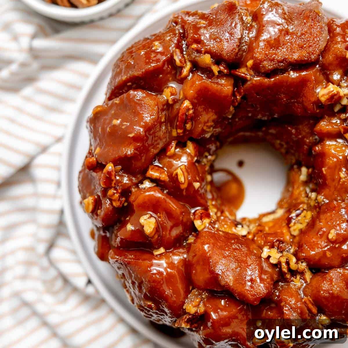Cinnamon Pull Apart Monkey Bread with Biscuits 11 A close-up of a glazed monkey bread ring with pecans on a white plate, set on a striped cloth.