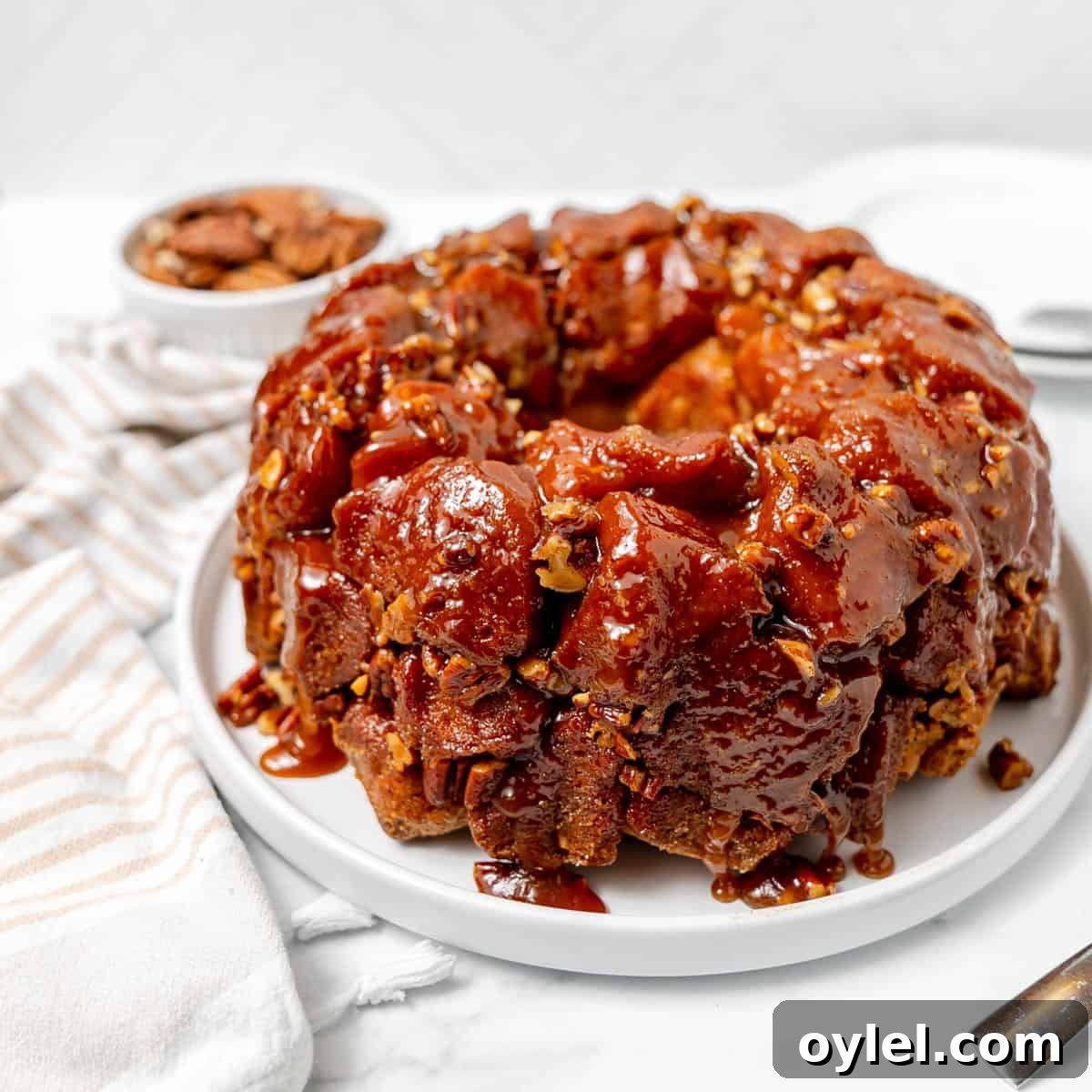 Cinnamon Pull Apart Monkey Bread with Biscuits 12 A round glazed monkey bread with pecans sits on a white plate, with a striped napkin and a small bowl of pecans in the background.