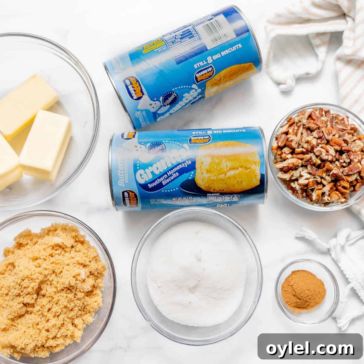 Cinnamon Pull Apart Monkey Bread with Biscuits 3 Two cans of biscuit dough, bowls of butter, brown sugar, white sugar, chopped pecans, and ground cinnamon are arranged on a white surface with a striped towel.