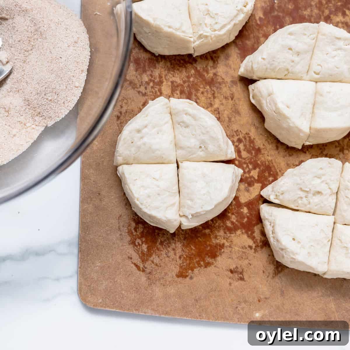 Cinnamon Pull Apart Monkey Bread with Biscuits 5 Quartered biscuit dough pieces are arranged on a wooden cutting board next to a glass bowl containing a cinnamon-sugar mixture.