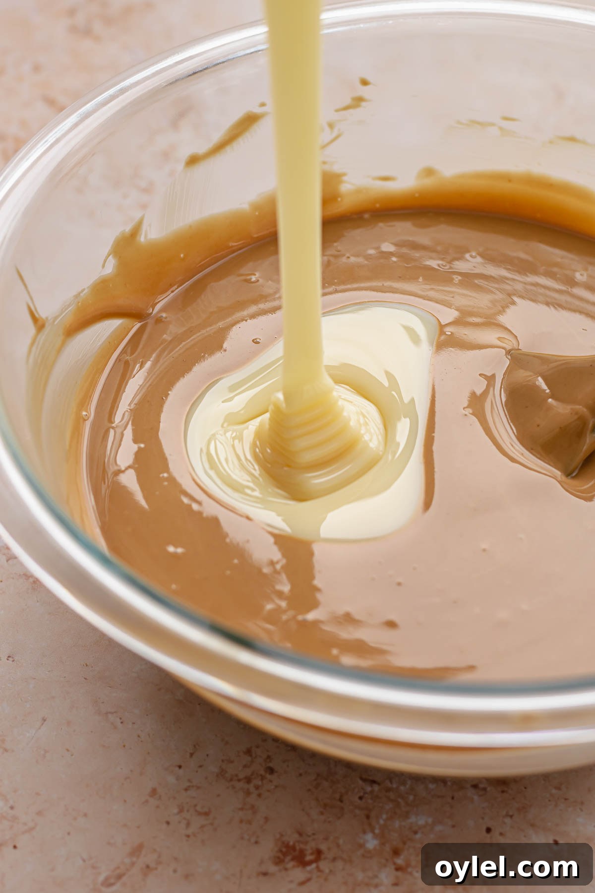 Sweetened condensed milk pouring into a bowl of white chocolate and cookie butter.